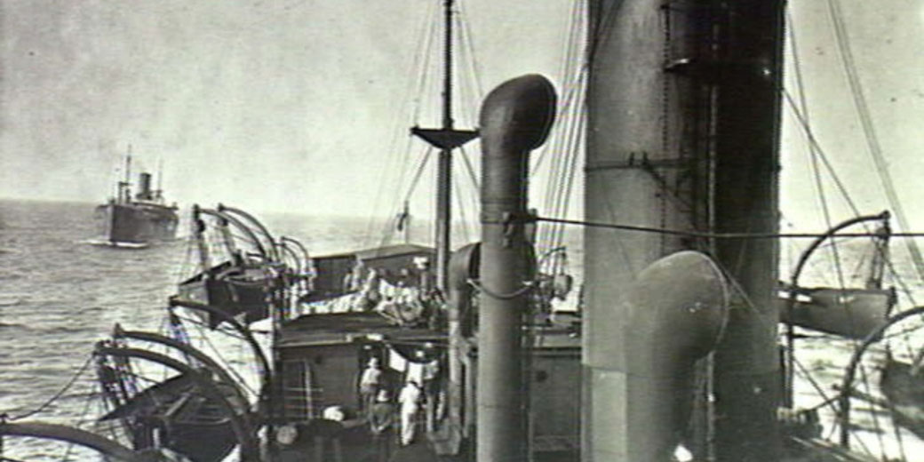 View from a ship’s deck showing lifeboats, crew members, and another vessel following behind on open water.