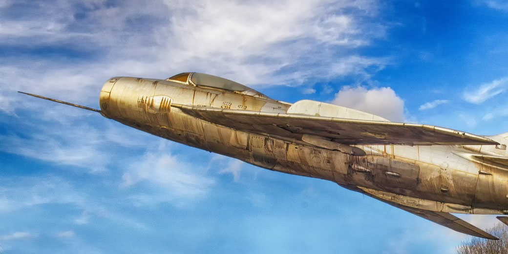 Close-up of an old, weathered military jet angled upward against a blue sky, appearing mounted for display rather than in flight.