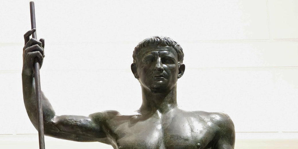 Bronze statue of a muscular man holding a spear upright, with a stern expression and short-cropped hair, symbolizing strength and authority.
