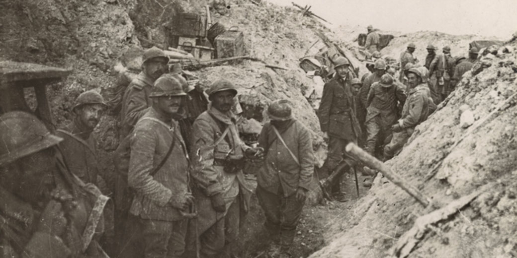 Soldiers in helmets stand and rest in a muddy World War I trench, surrounded by rough terrain and scattered supplies.