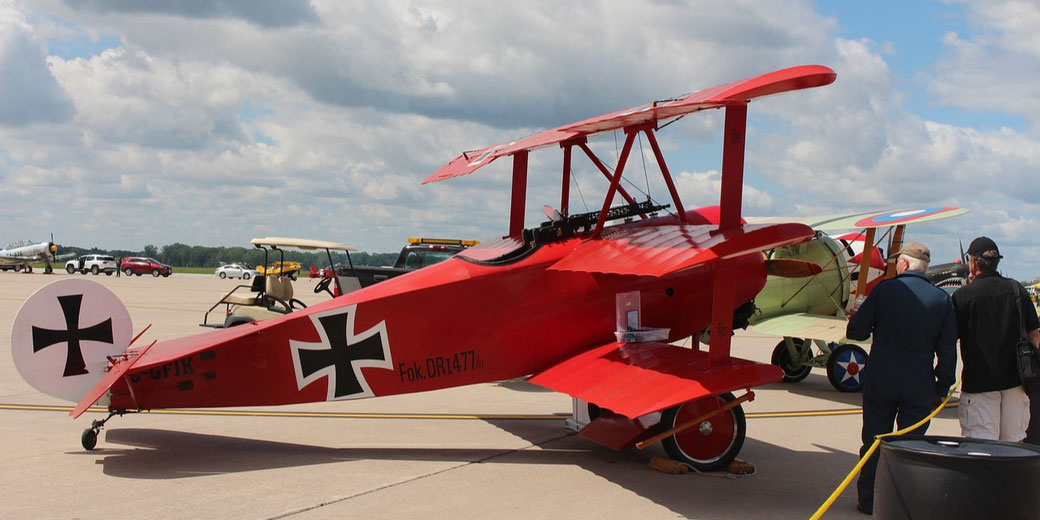 A bright red Fokker Dr.I triplane replica is on display at an airfield, with people observing it up close.