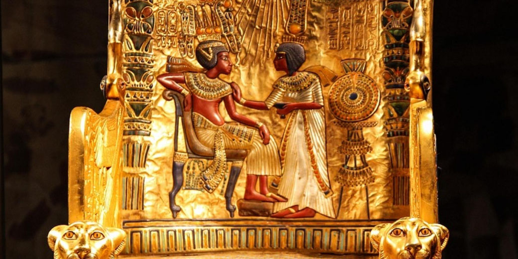 Gold-covered ancient Egyptian throne with painted figures, hieroglyphs, and lion-head armrests, lit to highlight detailed craftsmanship.