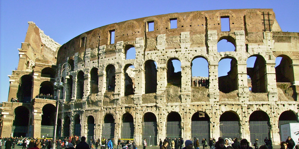 The Colosseum in Rome, highlighting its partially damaged exterior with arched openings and remnants of its ancient structure, surrounded by tourists under a clear blue sky.