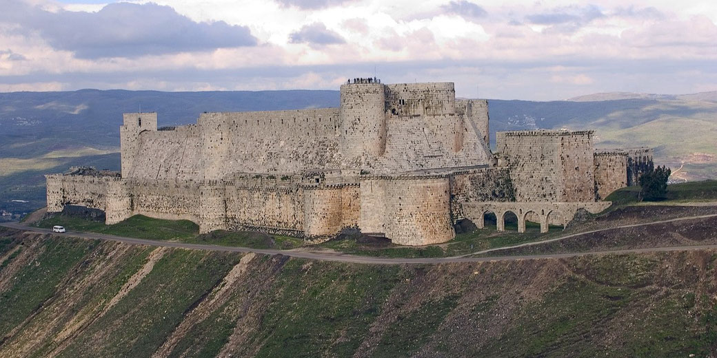 A view of Krak des Chevaliers, a Crusader castle in Syria. The fortress is perched on a hill, with its massive stone walls and towers.