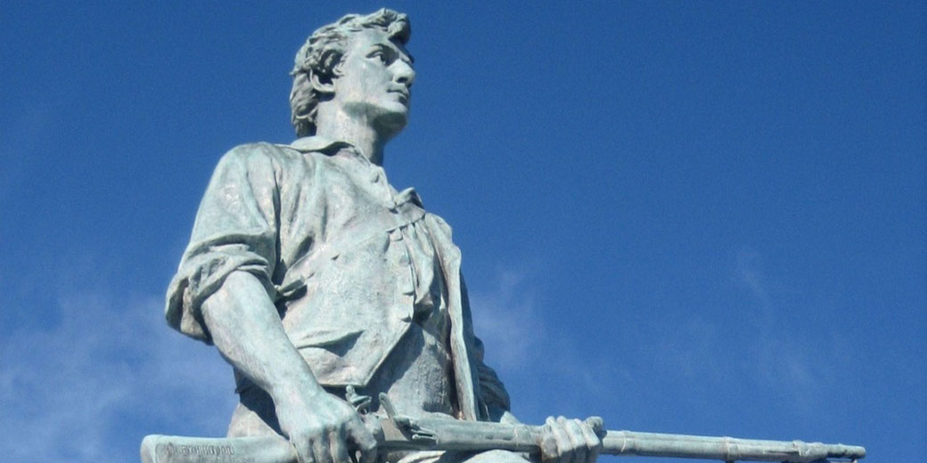 Bronze statue of a Revolutionary War soldier holding a musket, gazing forward with determination under a clear blue sky.