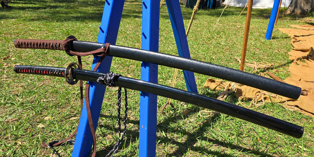 Two Japanese swords, a katana and a wakizashi, rest on a blue wooden stand. The top sword has a brown-wrapped handle, while the bottom has a black handle with red accents.