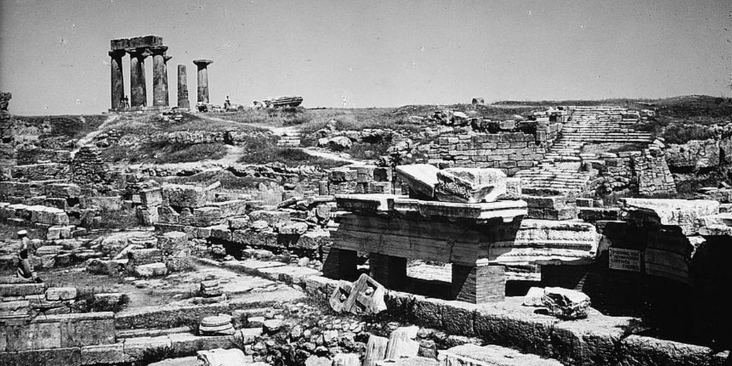 Black and white image of ancient ruins with broken columns, stone foundations, and steps scattered across a grassy hill.