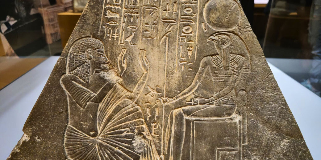 An ancient Egyptian carving depicting a worshipper with raised hands before Ra-Horakhty, a falcon-headed god with a sun disk. Hieroglyphs surround the figures on a triangular stone slab.