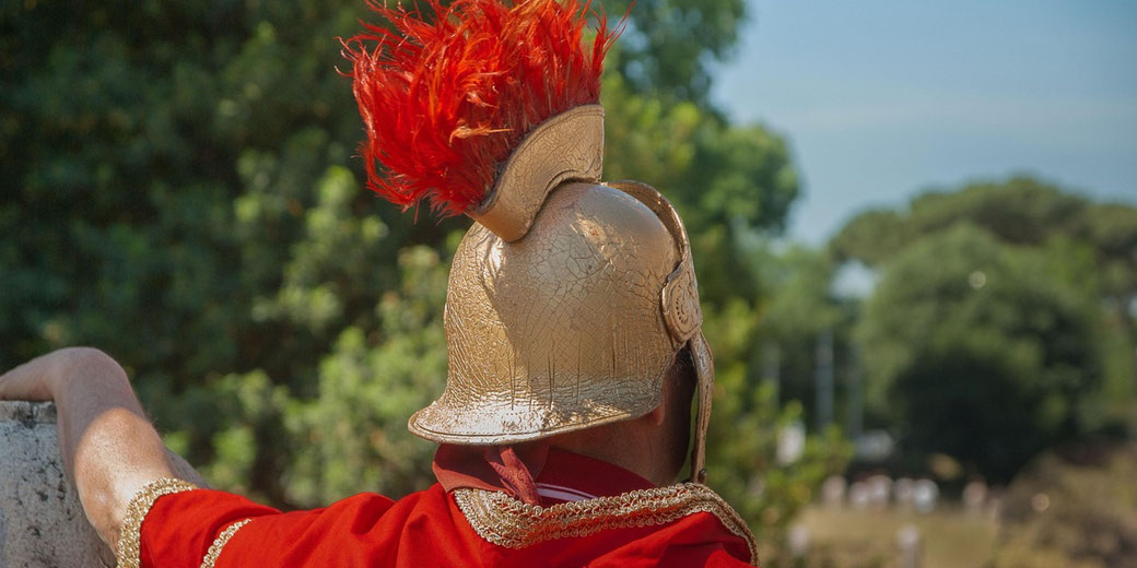 A Roman reenactor from behind, wearing a gold helmet with a red plume and a red cloak. He gazes into the distance.