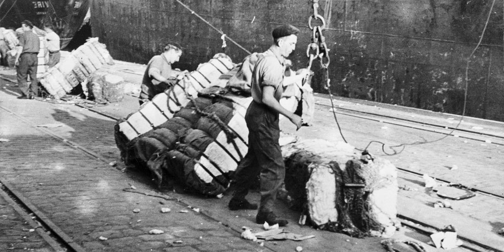 Black and white photo of dock workers loading large bales onto a ship using chains and hooks at an industrial port.