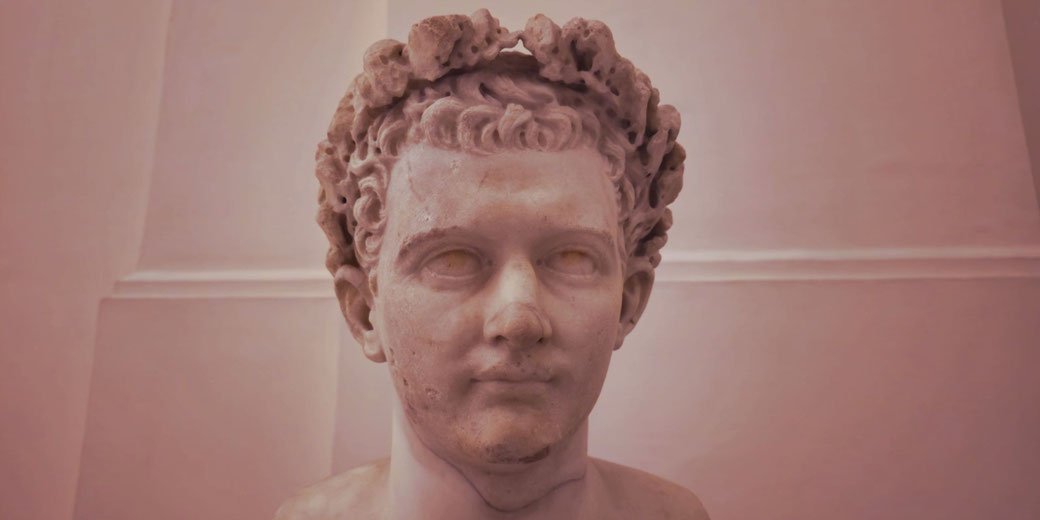 Marble bust of a young man with short curly hair wearing a laurel wreath, eyes blank, expression serene.