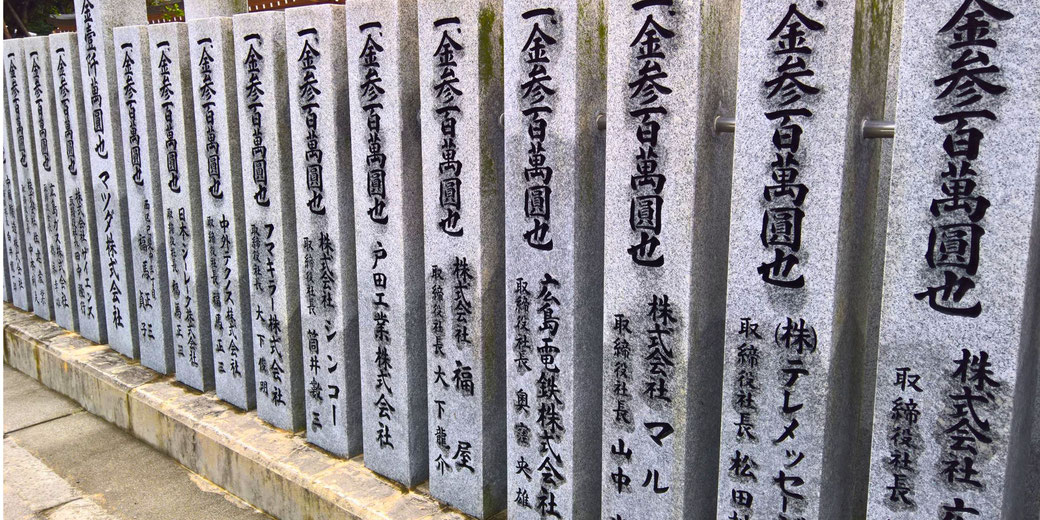 A row of stone pillars with Japanese inscriptions, likely representing donor names and contributions to a temple or shrine. The black calligraphy contrasts with the light grey stone.