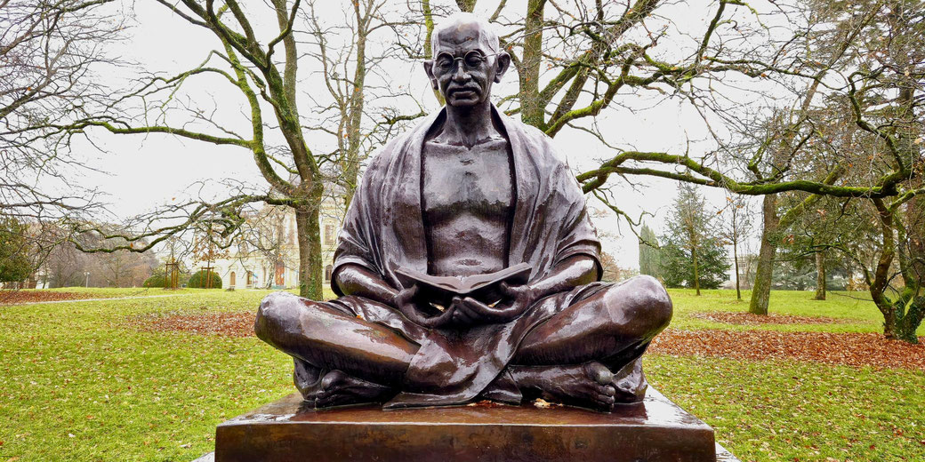 A bronze statue of Mahatma Gandhi seated in a meditative pose, holding an open book. Set in a park with bare trees, it evokes a sense of peace, wisdom, and contemplation.