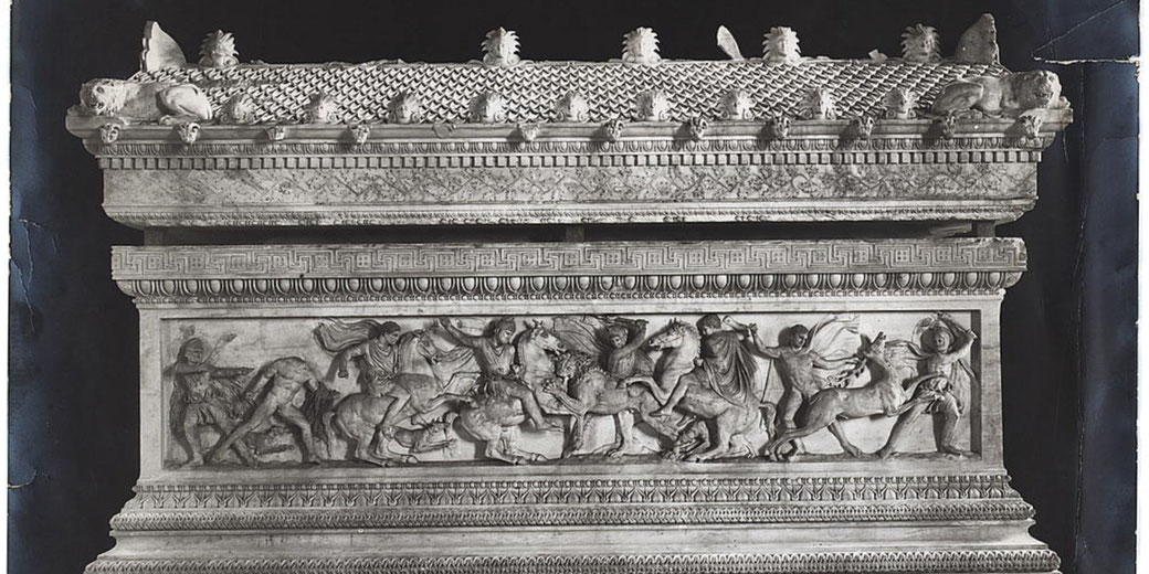 Ancient marble sarcophagus with detailed reliefs of warriors and horses in battle, topped with lion sculptures and decorative patterns.