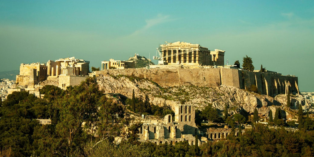 Ancient Acropolis in Athens with the Parthenon atop a rocky hill, surrounded by ruins and greenery under a clear sky.