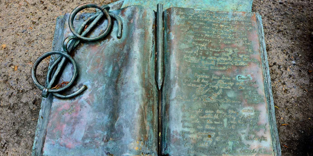 An open metal book sculpture with large ring bindings and a pen down the center, engraved text on the right page, resting on a gravel surface outdoors.