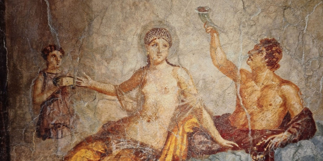 Ancient fresco depicts a semi-nude woman seated between a man raising a drinking horn and a servant girl holding a cup, suggesting a banquet or mythological scene.