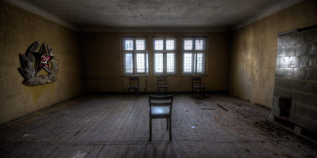 Dimly lit abandoned room with five wooden chairs, peeling walls, Soviet emblem on the left wall, and a tiled stove on the right.