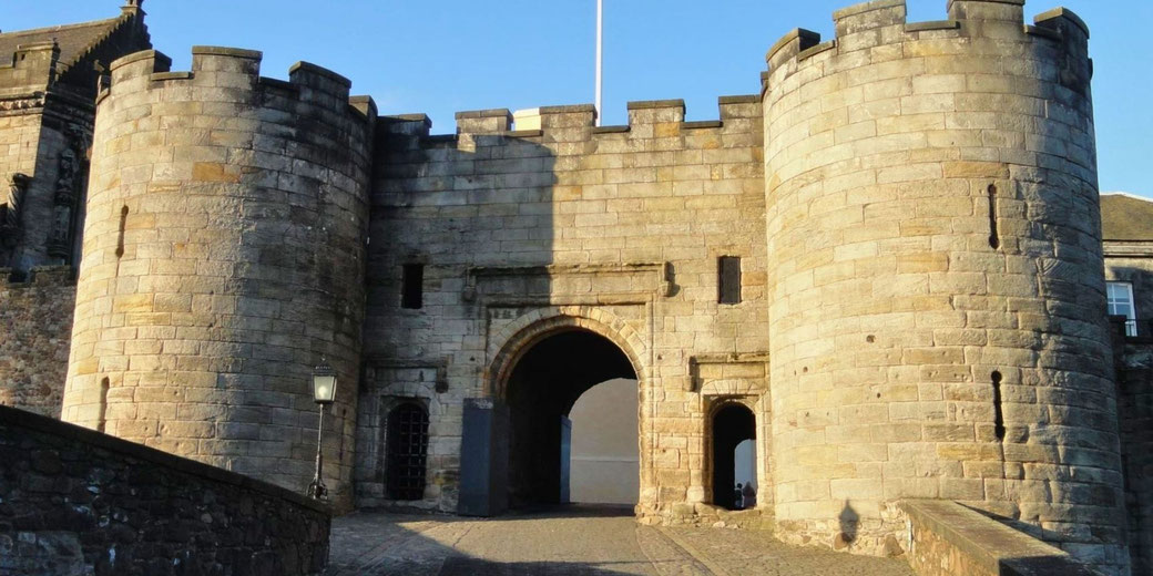 The fortified entrance of Stirling Castle, featuring large round stone towers flanking an arched gateway. The medieval structure is bathed in sunlight, highlighting its imposing presence.