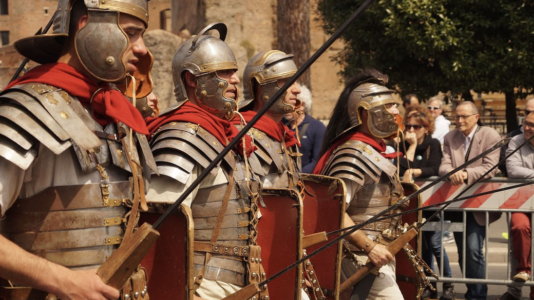 Group of reenactors dressed as Roman legionaries with armor, helmets, red cloaks, and shields marching in front of a crowd.