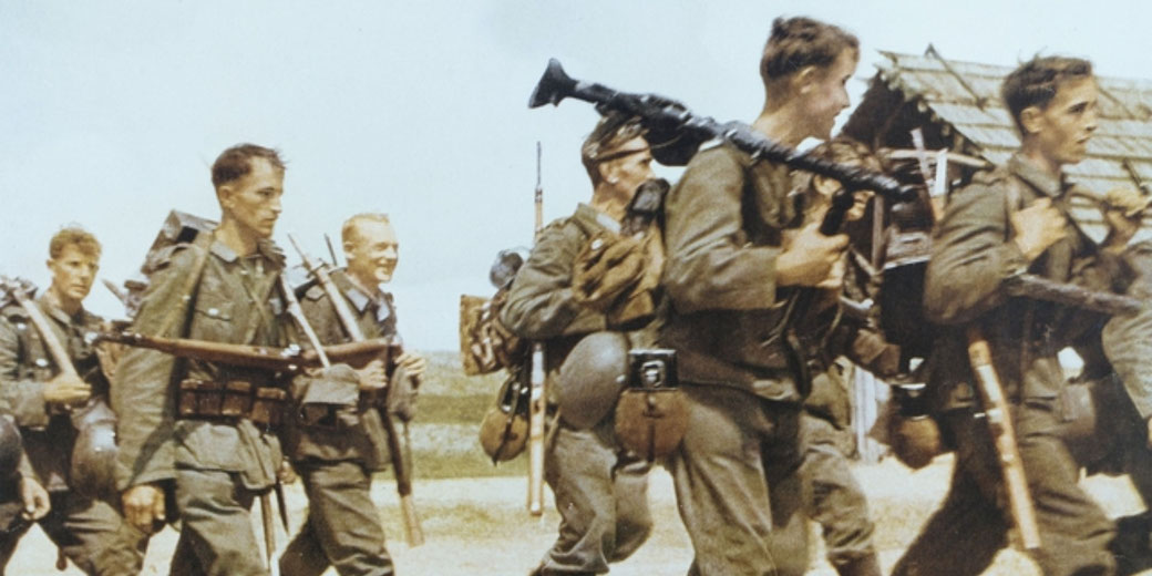 A group of soldiers marches in uniform, carrying rifles and gear, passing by a rustic wooden structure under a clear sky.