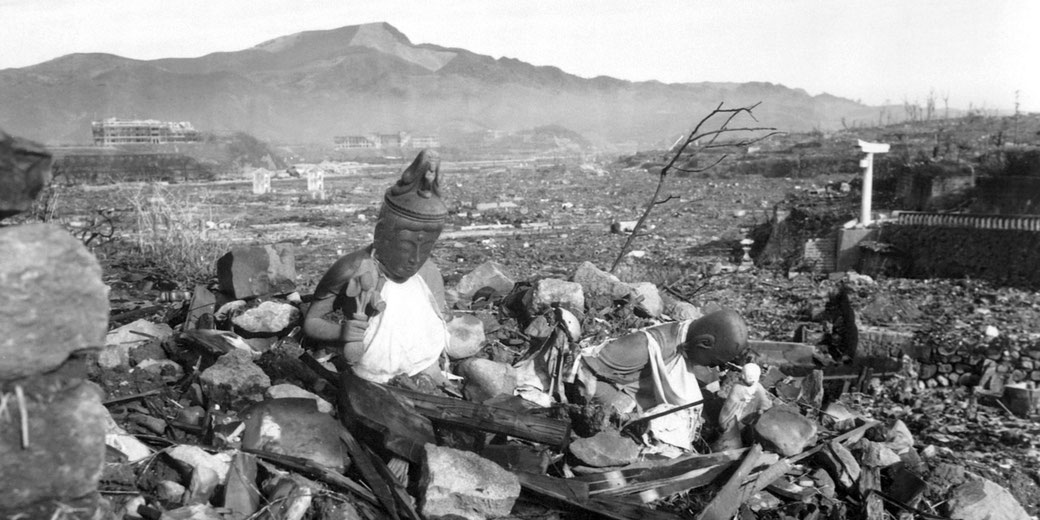 Buddhist statues sit amid rubble in Nagasaki after the atomic bombing, surrounded by widespread destruction and distant mountains.