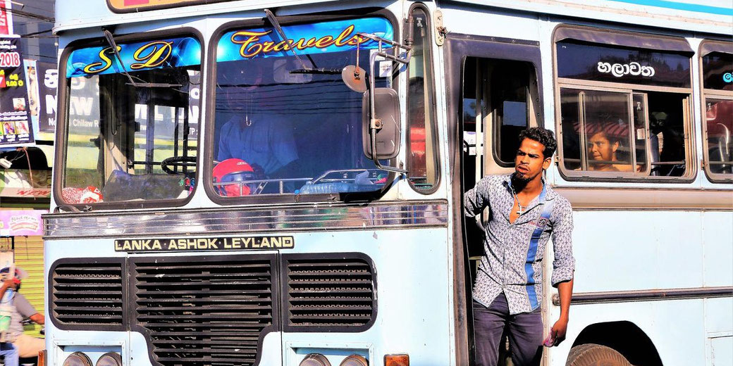 A man stands at the entrance of a blue public bus labeled "Lanka Ashok Leyland" with passengers visible inside, including a woman by the window.