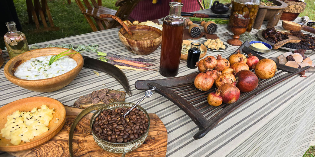 A table laden with historical-style food, including pomegranates, dips, carrots, spices, pickled items, and bottles of drink. The setup features rustic bowls, utensils, and a striped cloth backdrop.