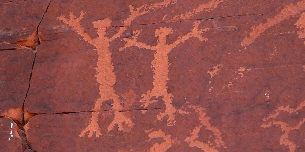 Ancient petroglyphs on reddish rock, depicting two humanoid figures with raised arms. The carvings are weathered, likely created by early inhabitants as part of symbolic or ritual art.