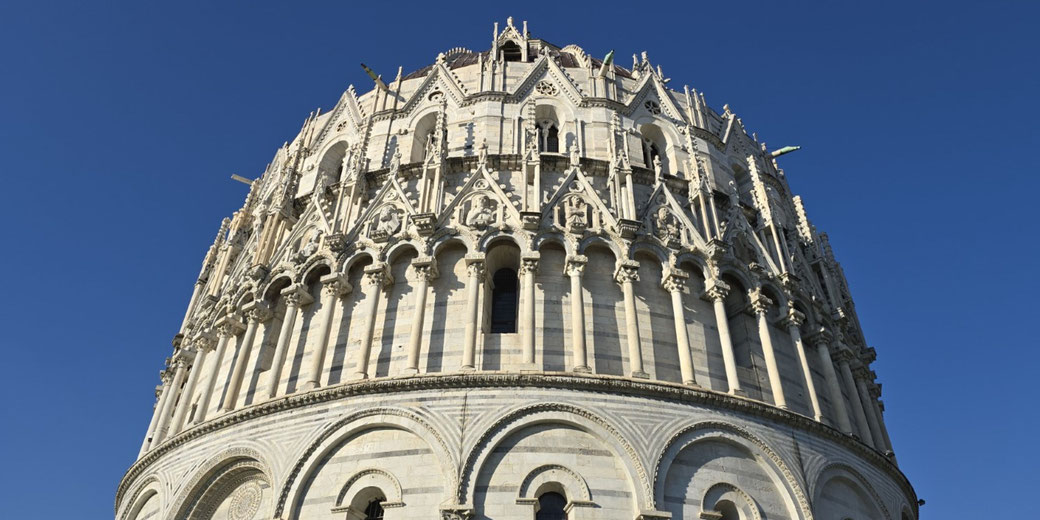 The upper section of the Pisa Baptistery, featuring ornate Gothic architecture, arched columns, and detailed stone carvings against a clear blue sky.
