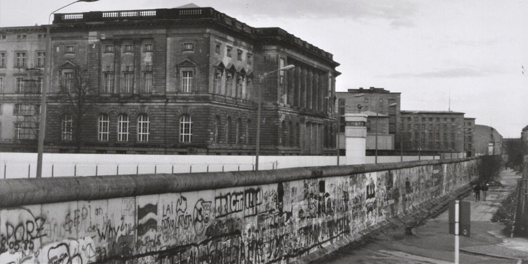 Black and white photo of the Berlin Wall covered in graffiti, with guard towers and old buildings in the background.