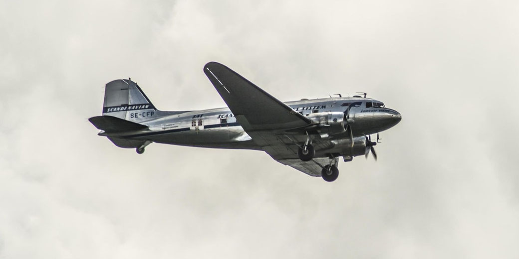 Vintage twin engine propeller aircraft with Scandinavian Airlines markings flying with landing gear down against a cloudy sky, side view of polished metal fuselage in flight.