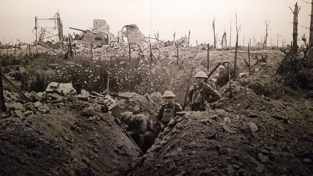 Three soldiers in a trench surrounded by rubble and barbed wire, with destroyed buildings and shattered trees in the background during World War I.
