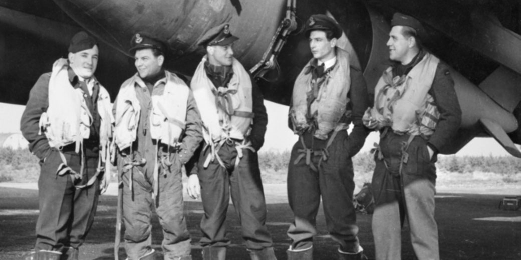 Five WWII airmen in flight suits and life vests stand together beneath an aircraft, chatting and smiling.