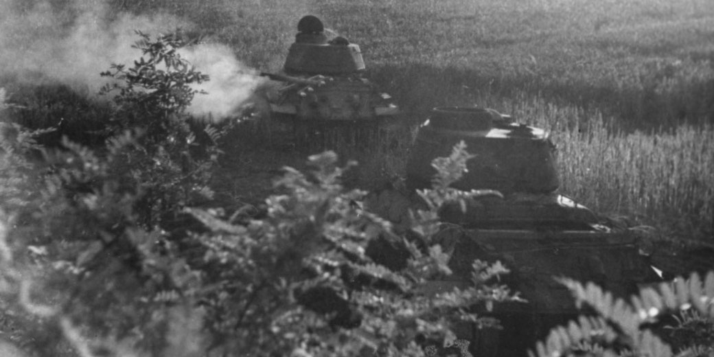 Black and white wartime photo of tanks advancing through tall grass while firing. Smoke drifts across the field as armored vehicles move in formation during a Korean War battlefield advance.