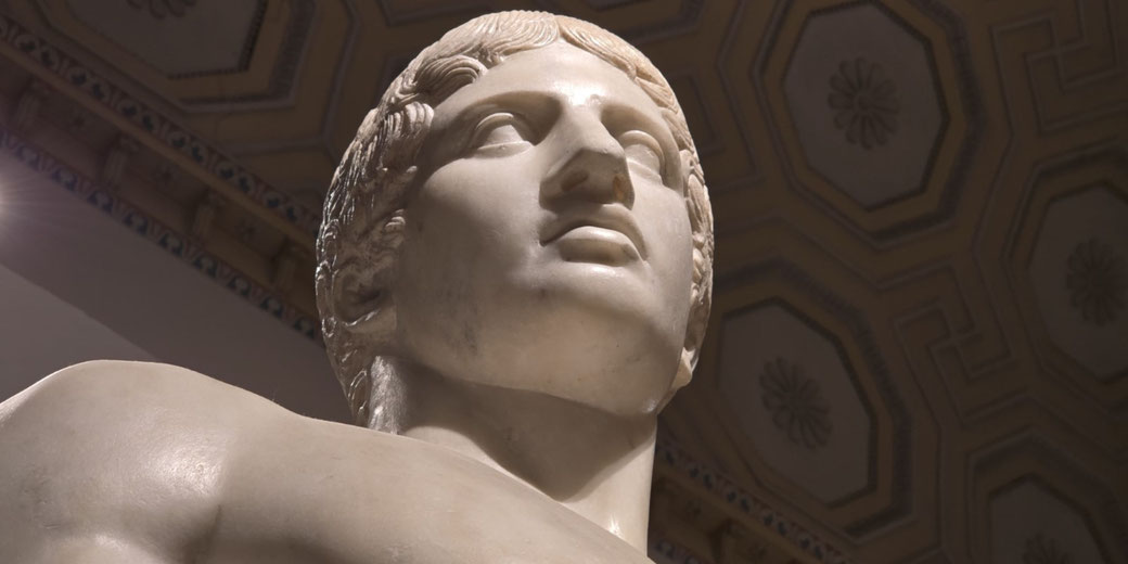 Close-up of a marble statue’s head and upper torso, showing idealized facial features, wavy hair, and a strong neck, viewed from a low angle.