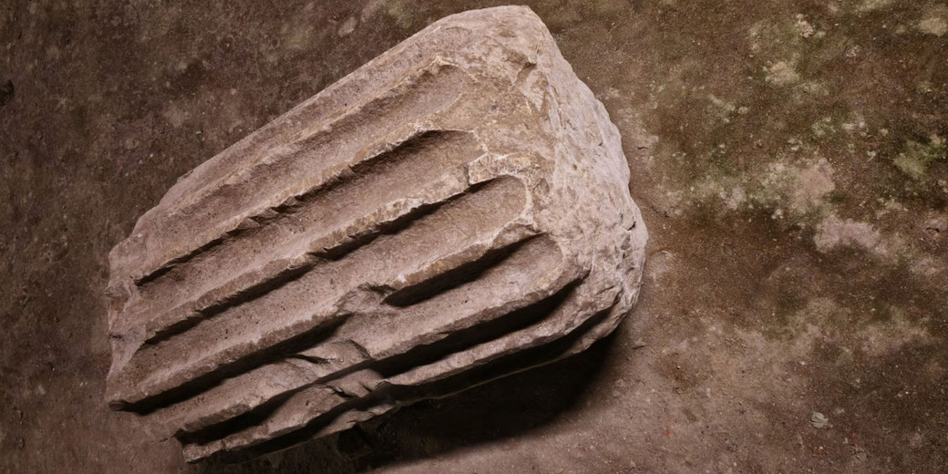 A broken fluted stone column segment lies on a rough, mottled ground surface, showing wear and erosion on its carved grooves.