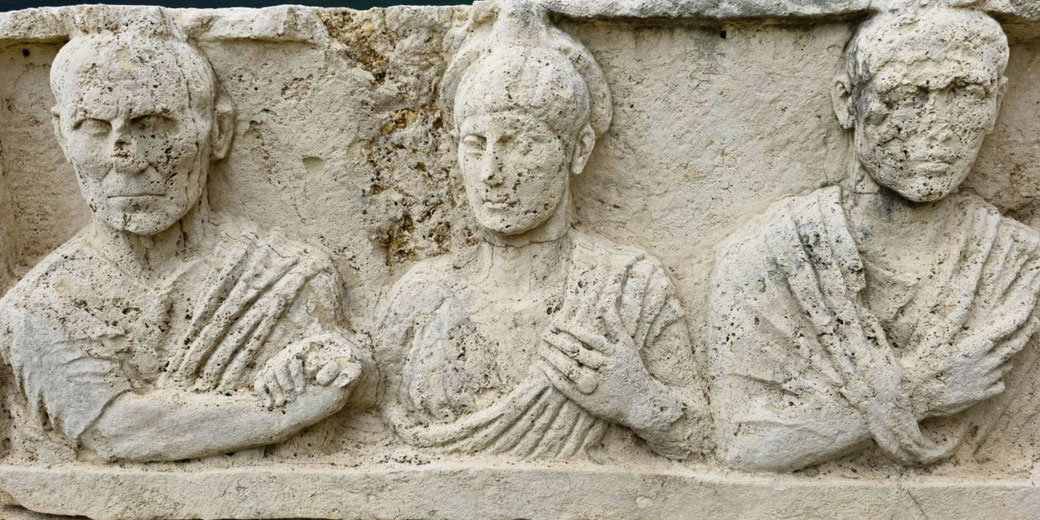 Weathered stone relief depicting three Roman figures in traditional attire, carved in a frontal, half-length pose.