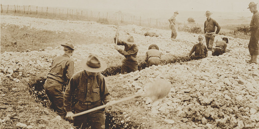 Soldiers dig and shape trenches in rocky terrain, some standing while others work with shovels under supervision during World War I.