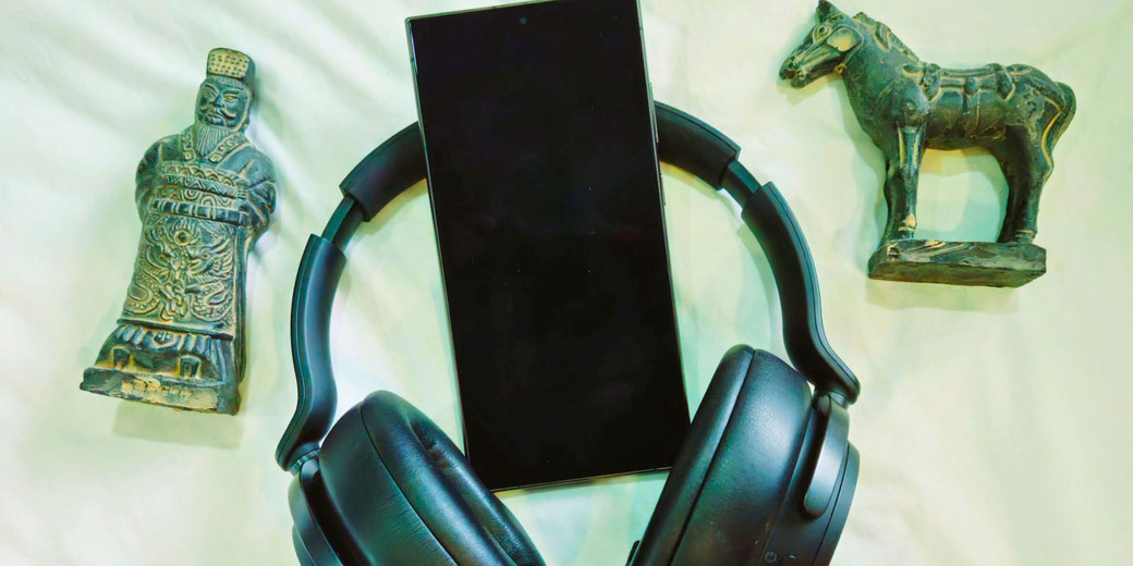 A smartphone, black headphones, a Chinese warrior figurine, and a horse statue on a white surface, symbolizing a blend of history and modern technology, possibly for history podcasts.
