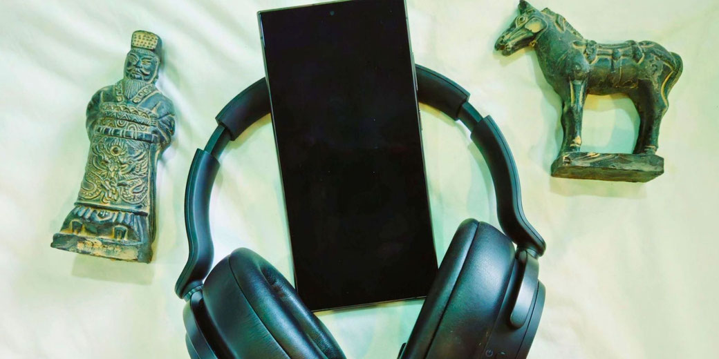 A smartphone, black headphones, a Chinese warrior figurine, and a horse statue on a white surface, symbolizing a blend of history and modern technology, possibly for history podcasts.