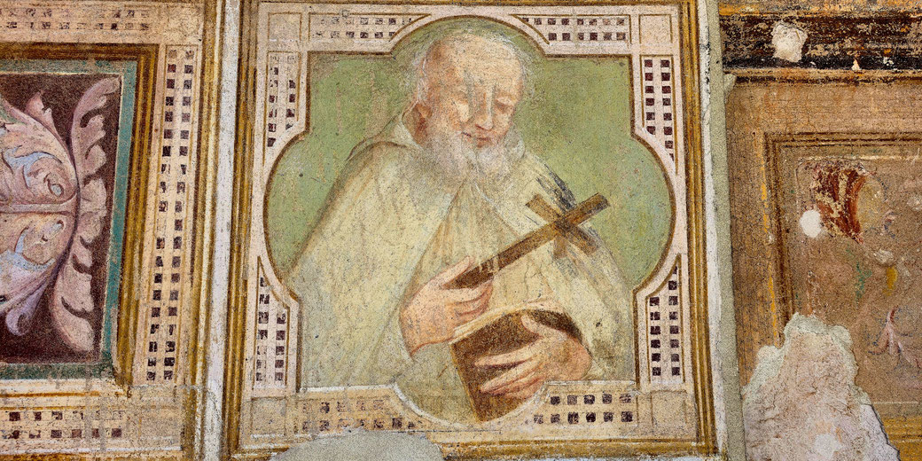 Fresco of an elderly monk with a white beard, holding a wooden cross and book, set within a decorative, bordered frame.