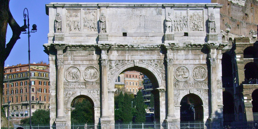Triumphal Roman arch with detailed reliefs and statues, standing in front of modern and historic buildings under a clear blue sky.