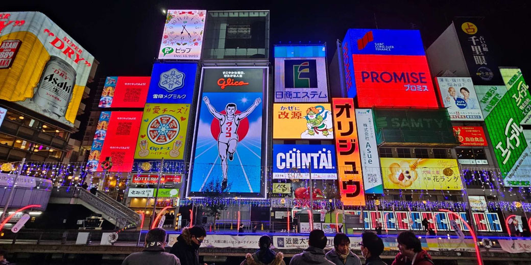 Night scene of Dotonbori with bright signs, canal frontage, and people gathered beneath the Glico runner billboard.