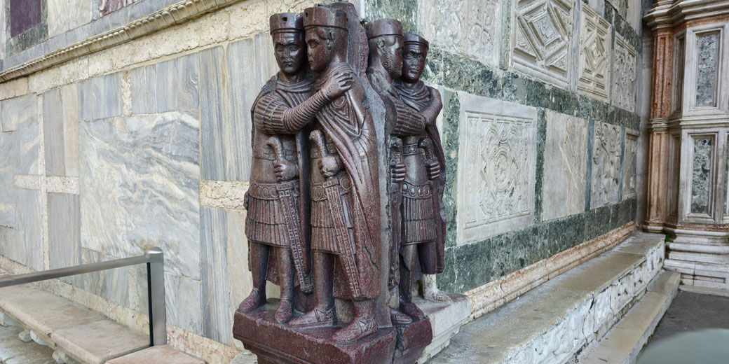 Four porphyry robed figures embracing at a basilica corner, set into banded marble and greenstone panels beside a water basin at St Mark’s Basilica in Venice.