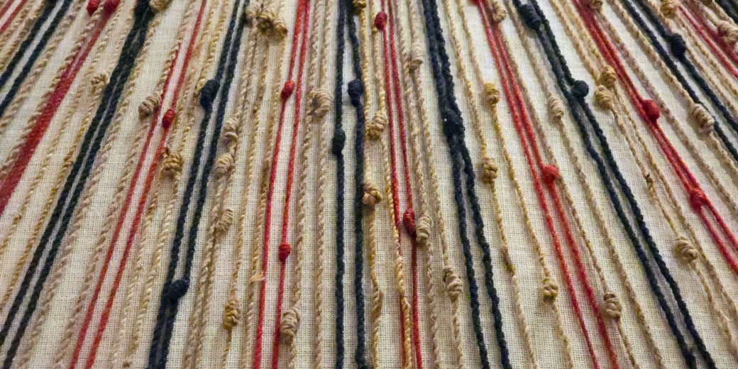 A close-up of textured fabric with vertical, multicolored yarn strands in red, black, and beige, some twisted and knotted, creating a raised, tactile pattern on a light-colored woven background.