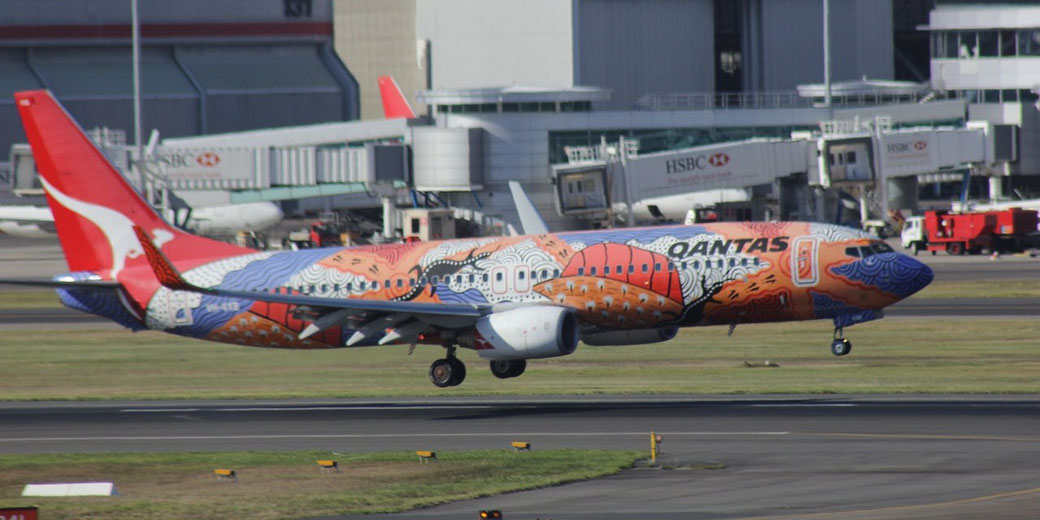 A Qantas airplane with bold Aboriginal art-style livery lands on a runway at a busy airport.