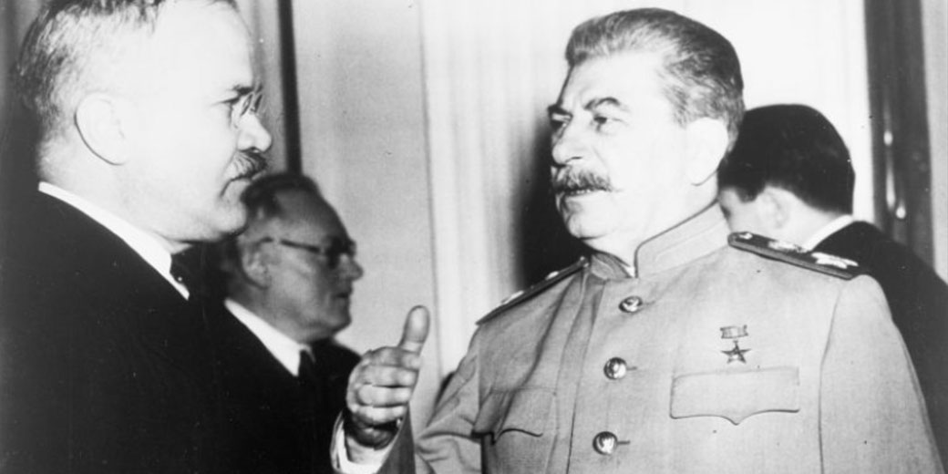 Black-and-white photo of Soviet leader Joseph Stalin speaking with another official in a formal setting, both appearing engaged in conversation.