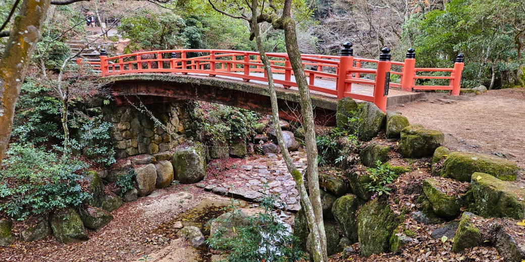 A red arched bridge with black finials spans a rocky, leaf-covered stream bed in a wooded area with moss-covered stones and trees.