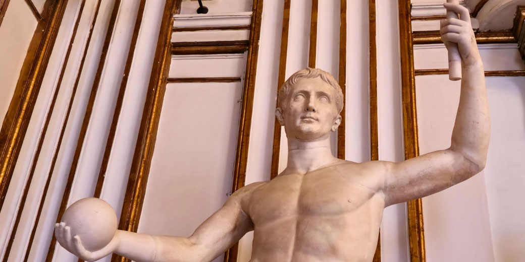 Marble statue of a man with a scroll in one hand and a sphere in the other, standing against a decorated wall with gold-accented vertical panels.